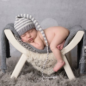 May include: A newborn baby wearing a gray and white striped knit hat with a long tail sleeps on a white wooden prop with a gray knitted blanket. The prop is on a gray fluffy surface.
