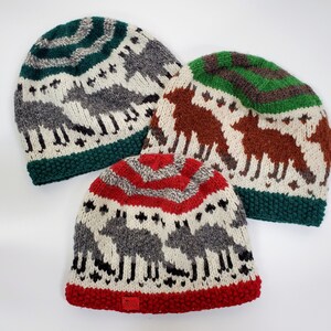 Könnte beinhalten: Drei gestrickte Wintermützen mit weißem Hintergrund und einem Muster aus grauen Tieren. Die Mützen haben unterschiedlich farbige Kanten: grün, braun und rot.
