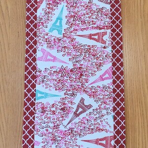 May include: A red and white table runner with a pattern of Eiffel Towers and hearts. The runner has a burgundy border with a geometric pattern.