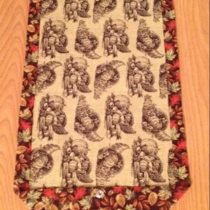 May include: A brown and beige table runner with a fall theme. The runner features a repeating pattern of cornucopias filled with fruit and vegetables. The edges of the runner are trimmed with a brown, green, and red leaf pattern.