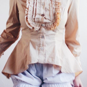 Peut inclure: Une blouse marron clair avec un col à volants et des manches longues. La blouse a un corsage ajusté et une jupe ample. La blouse est portée sur une paire de bloomers blancs avec des bordures en dentelle.