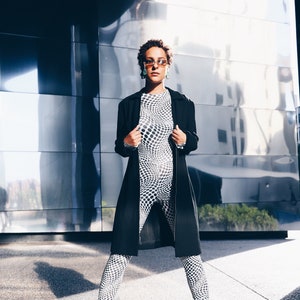May include: A woman wearing a black coat over a white and black patterned jumpsuit. She is wearing sunglasses and earrings. The woman is standing in front of a reflective wall.