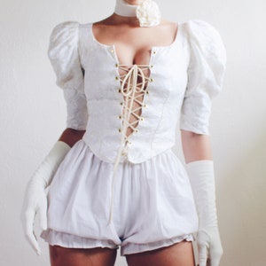May include: White lace-up top with puffed sleeves and a white, ruffled,  short skirt. The top has a low neckline and is tied at the front with a white ribbon.