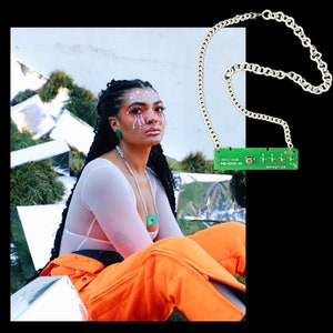 May include: A woman wearing a white top and orange pants with a green circuit board necklace. The necklace has a silver chain and a green circuit board with the text "DISPLAY BOARD 640-0245C-03" and a red button.