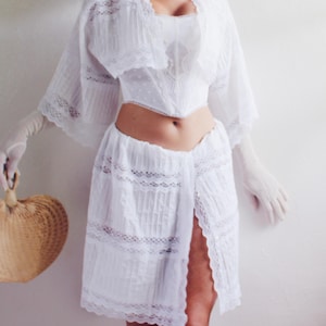 Pu&ograve; includere: Abito bianco in pizzo con una gonna lunga e fluida e un corpetto aderente. L'abito ha una spacco anteriore e un bordo in pizzo.