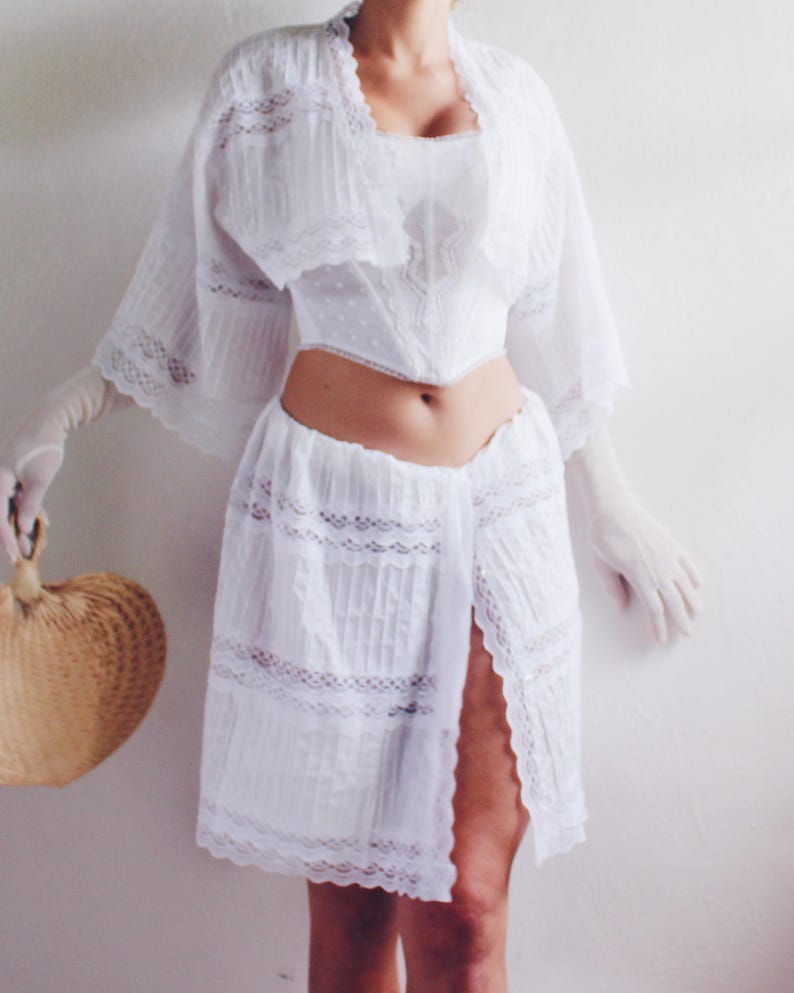 Pu&ograve; includere: Abito bianco in pizzo con una gonna lunga e fluida e un corpetto aderente. L'abito ha una spacco anteriore e un bordo in pizzo.