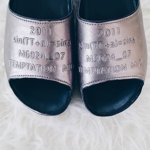 Może przedstawiać: Para metalicznych, srebrnych sandałów z otwartymi palcami. Sandały mają ciemną podeszwę i wytłoczony tekst: "2011", "sin(TT+a)=sina", "M6024_07" i "TEMPTATION MIO". Na białym, puszystym tle.