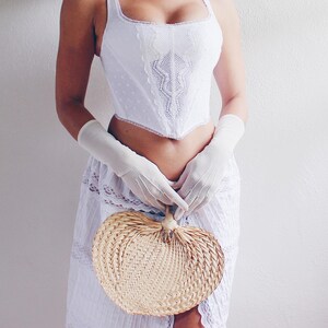 Pu&ograve; includere: Un corpetto bianco in pizzo con scollo a cuore e una gonna bianca con bordo in pizzo. La persona tiene un ventaglio intrecciato a forma di cuore.