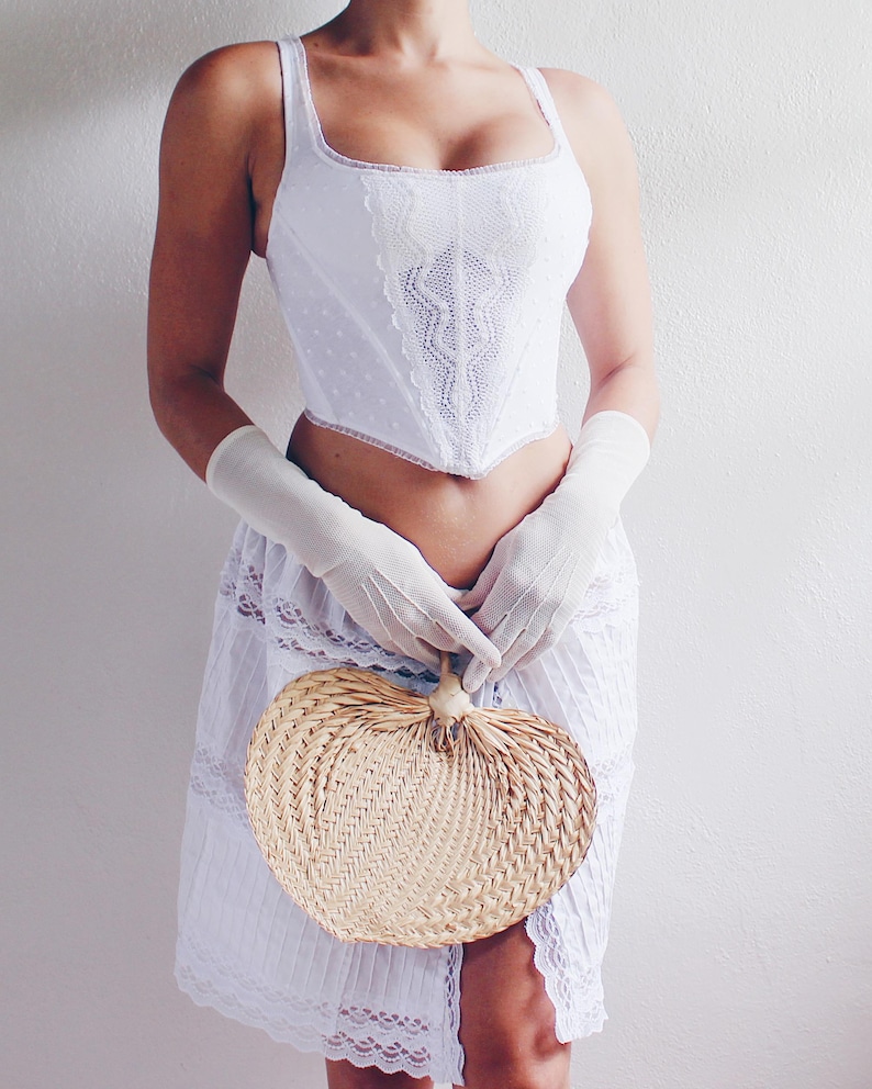 Pu&ograve; includere: Un corpetto bianco in pizzo con scollo a cuore e una gonna bianca con bordo in pizzo. La persona tiene un ventaglio intrecciato a forma di cuore.