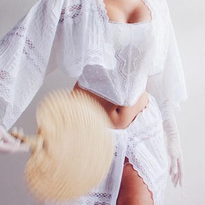 Pu&ograve; includere: Un top di pizzo bianco con un corsetto di pizzo bianco e una gonna di pizzo bianco. La persona tiene un ventaglio bianco.