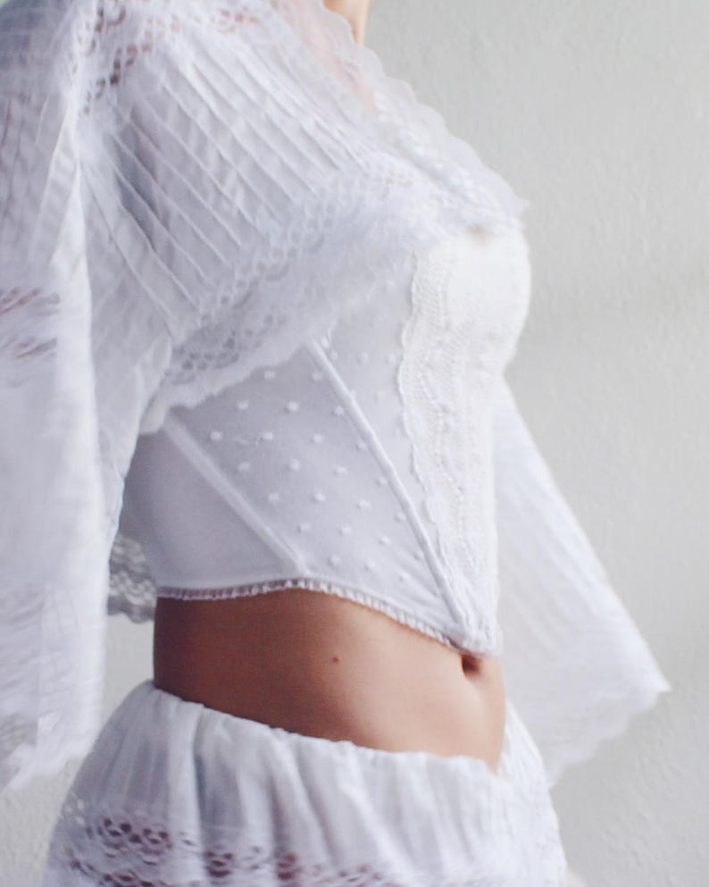 Pu&ograve; includere: Un top bianco in pizzo con un corpetto aderente e una gonna fluida. Il top presenta un delicato motivo floreale e una scollatura a cuore.