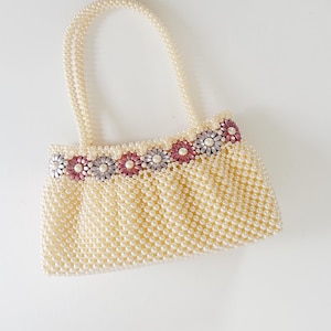 Pode incluir: Bolsa de mão de contas de cor marfim com um design floral. A bolsa tem formato retangular com duas alças e uma faixa decorativa de flores rosa, prateadas e lavanda na parte superior. A bolsa é feita de pequenas contas redondas.