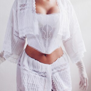 Pu&ograve; includere: Un top bianco in pizzo con un corpetto aderente e un'sovrapposizione fluida e trasparente. Il top ha una scollatura a cuore e maniche lunghe con bordi in pizzo. La modella indossa una gonna bianca con bordi in pizzo.