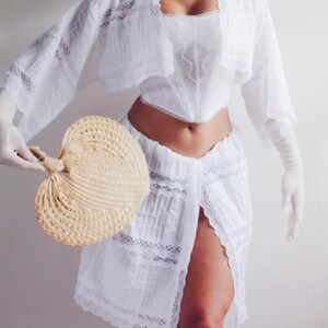 Pu&ograve; includere: Un set di top e gonna in pizzo bianco con un orlo arricciato e un bordo in pizzo. Il top ha una scollatura a cuore e un corpetto aderente. La gonna &egrave; a trapezio e ha uno spacco sul davanti. La modella tiene un ventaglio intrecciato.