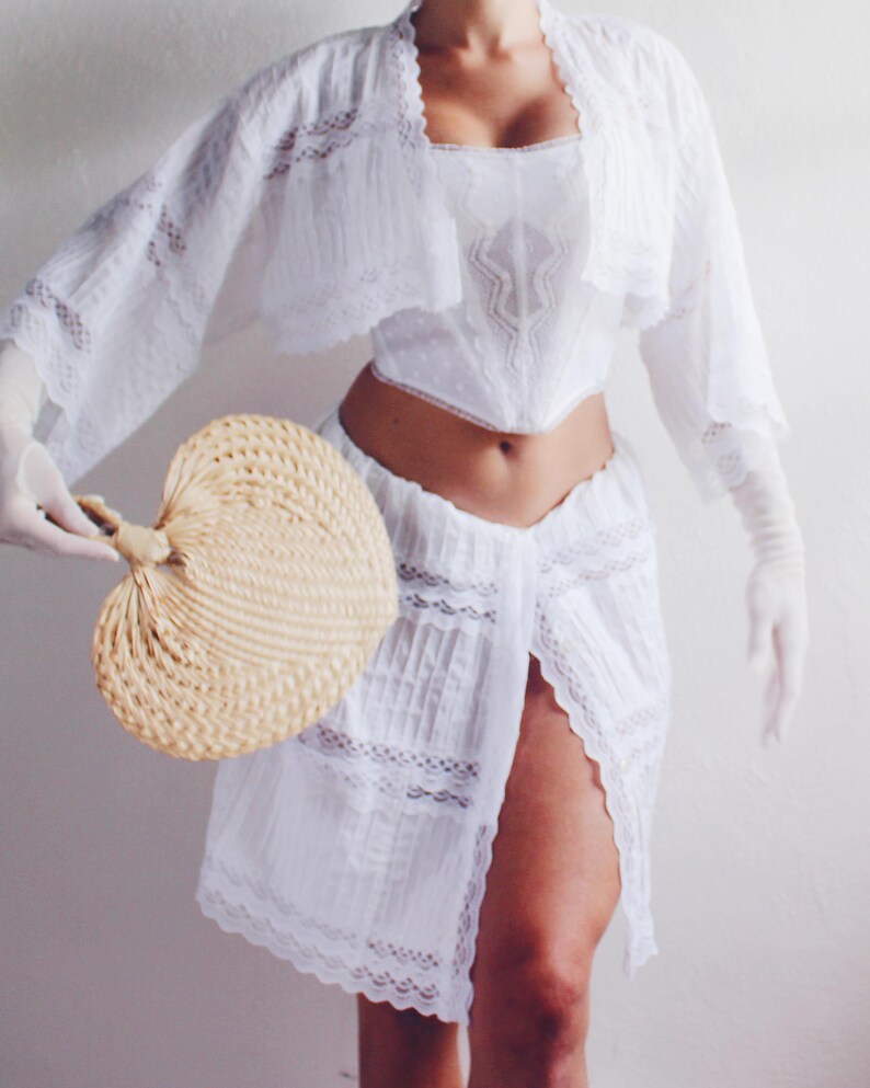 Pu&ograve; includere: Un set di top e gonna in pizzo bianco con un orlo arricciato e un bordo in pizzo. Il top ha una scollatura a cuore e un corpetto aderente. La gonna &egrave; a trapezio e ha uno spacco sul davanti. La modella tiene un ventaglio intrecciato.