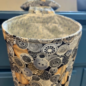 May include: A fabric hanging trash bag with a button pattern in shades of gray, tan, and black. The interior lining features a small floral pattern. The bag has a loop for hanging.