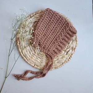 May include: A brown crocheted baby bonnet with ties. The bonnet is sitting on a woven straw mat.