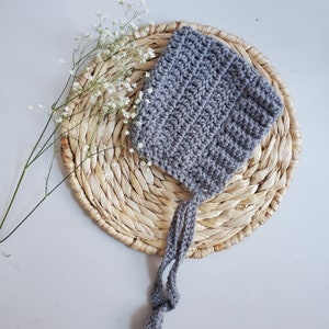 May include: A gray crocheted baby bonnet with a long tie. The bonnet is sitting on a woven straw mat.