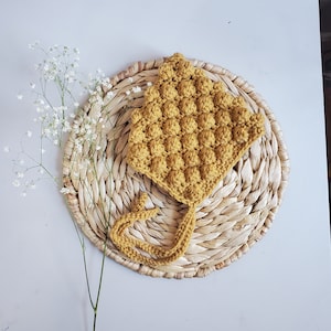 May include: A crocheted baby bonnet in a mustard yellow color with a textured, bubble-like pattern. The bonnet is tied with a matching ribbon.