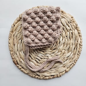 May include: A light brown crocheted baby bonnet with a textured bobble stitch pattern. The bonnet is tied with a long, thin ribbon.