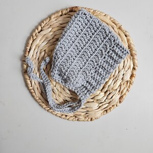 May include: A gray crocheted baby bonnet with ties. The bonnet is sitting on a woven straw round placemat.