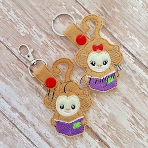 Może przedstawiać: Dwa brązowe breloczki z małpami z filcu z czerwonymi guzikami i fioletowymi książkami. Małpy trzymają książki i mają czerwoną kokardkę i białe oczy.