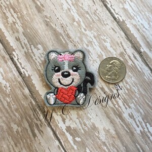 Puede incluir: Un parche bordado con un animal de dibujos animados que sostiene un coraz&oacute;n rojo. El animal es gris con un lazo rosa en la cabeza y est&aacute; sentado. El parche es aproximadamente del tama&ntilde;o de una moneda de euro.