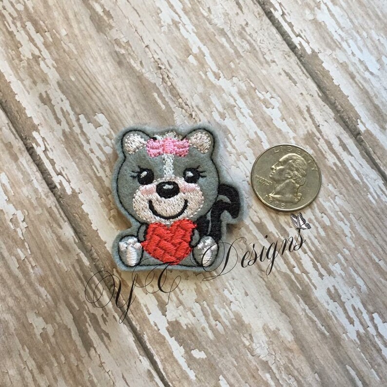 Puede incluir: Un parche bordado con un animal de dibujos animados que sostiene un coraz&oacute;n rojo. El animal es gris con un lazo rosa en la cabeza y est&aacute; sentado. El parche es aproximadamente del tama&ntilde;o de una moneda de euro.
