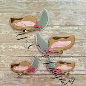 May include: Four pairs of felt hair clips with brown and gray ears and pink bows. The ears are shaped like a dog's ears.