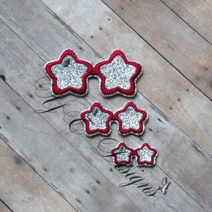 May include: Three pairs of star-shaped sunglasses with red frames and silver glitter lenses. The sunglasses are arranged in descending size on a wooden surface. The text "Designs" is visible in the lower left corner.