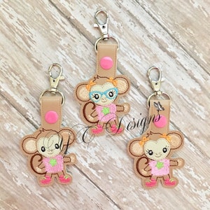 Może przedstawiać: Trzy haftowane breloczki z małpami w różowych i zielonych ubraniach. Małpy mają brązowe futro, różowe policzki i czarne oczy. Jedna małpa nosi okulary.