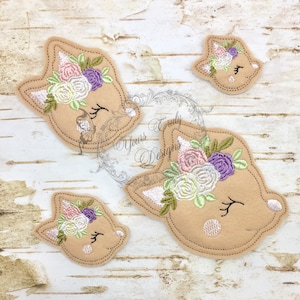 Puede incluir: Tres caras de cervatillo de fieltro bordadas con coronas de flores. La cara de cervatillo más grande es marrón con la nariz blanca y las orejas blancas. El cervatillo tiene un ojo cerrado y una sola pestaña. La corona de flores está hecha de flores rosas, blancas, moradas y verdes. La cara de cervatillo mediana es similar a la cara de cervatillo más grande, pero más pequeña. La cara de cervatillo más pequeña es similar a la cara de cervatillo mediana, pero aún más pequeña.