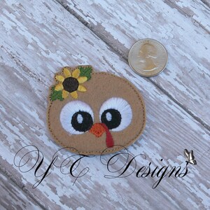 May include: Embroidered felt applique of a brown turkey with a yellow sunflower on its head. The turkey has large white eyes with black pupils and a red wattle.