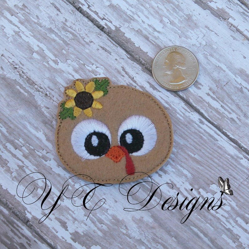 May include: Embroidered felt applique of a brown turkey with a yellow sunflower on its head. The turkey has large white eyes with black pupils and a red wattle.