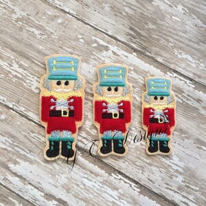 May include: Three felt nutcracker ornaments in different sizes. The largest nutcracker is red with a blue hat and a white face. The middle nutcracker is red with a blue hat and a white face. The smallest nutcracker is red with a blue hat and a white face.