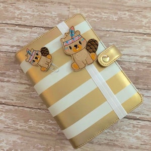 May include: A gold and white striped binder with a white elastic band. Two embroidered beaver bookmarks with colorful feathers are attached to the binder.