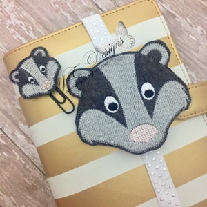 May include: Two embroidered badger faces, one large and one small, are attached to a gold and white striped notebook with a silver and white polka dot ribbon bookmark. The larger badger face is gray with a pink nose and white eyes. The smaller badger face is gray with a pink nose and white eyes. The notebook has the text "Designs" on the front.