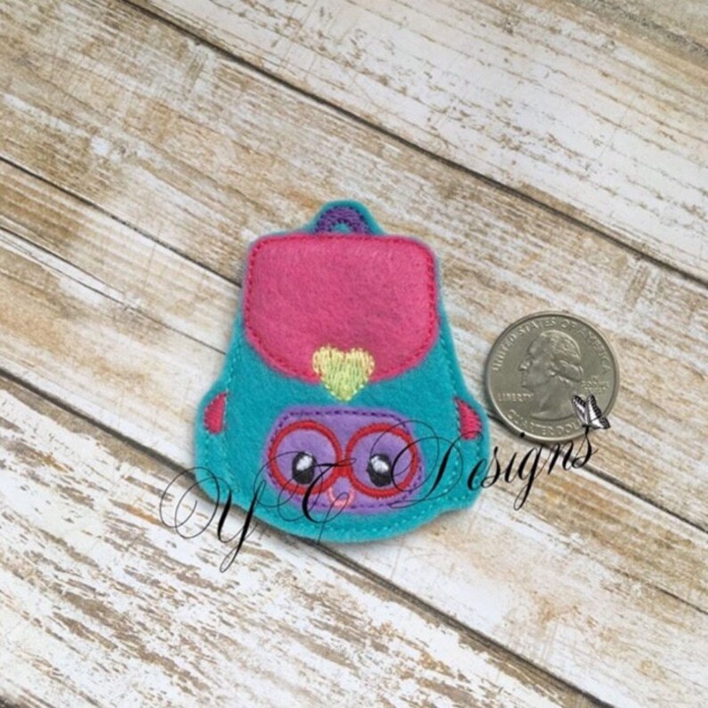 May include: A turquoise felt backpack with a pink flap, a yellow heart, and purple eyes. The backpack has a stitched design.