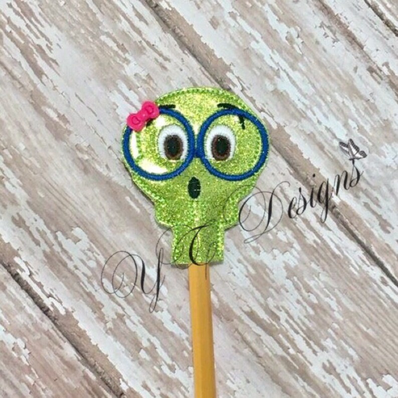 以下が含まれることがあります： 青い眼鏡とピンクのリボンをつけた漫画の顔をした緑色の鉛筆の飾り。飾りはキラキラした生地で作られています。