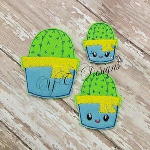 May include: Two felt cactus appliques with smiling faces. The cactus are green with yellow felt accents and are in blue pots.