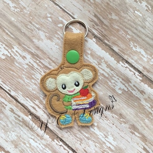 Może przedstawiać: Brelok do kluczy w kształcie brązowej małpy z filcu, trzymającej stos książek z jabłkiem na górze. Małpa ma na sobie niebieskie buty i zielony guzik na plecach.