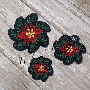 May include: Three embroidered felt patches featuring red poinsettia flowers with yellow centers and green leaves. The patches are on a woodgrain background.