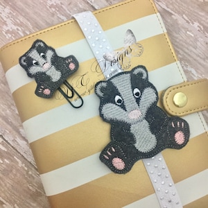 May include: Two gray and pink embroidered badger designs with white eyes. The larger badger is attached to a white ribbon with silver polka dots. The smaller badger is attached to a black binder clip. The designs are on a gold and white striped background.