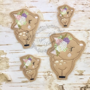 Puede incluir: Tres parches de cervatillo de fieltro bordados con coronas de flores. Los cervatillos son marrones con manchas blancas y tienen los ojos cerrados. Las flores son rosas, blancas, moradas y verdes.