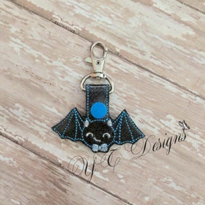 May include: A black bat-shaped keychain with a blue circle on its head and a gray bow tie. The keychain is attached to a silver metal key ring.