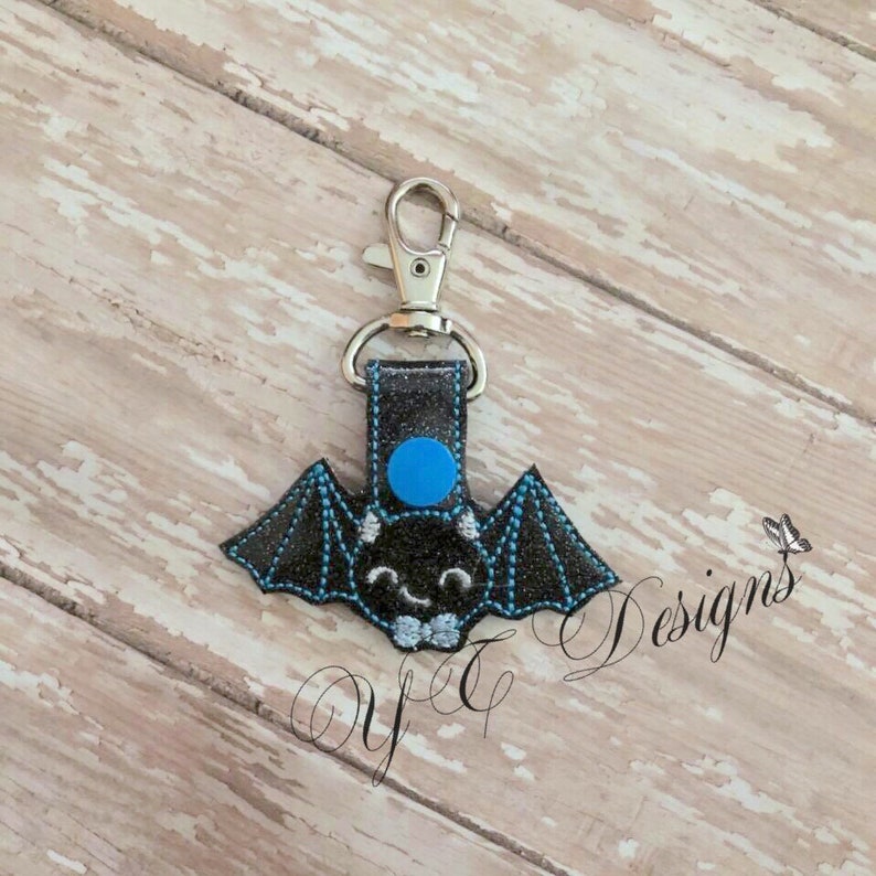 May include: A black bat-shaped keychain with a blue circle on its head and a gray bow tie. The keychain is attached to a silver metal key ring.