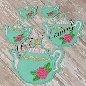 May include: Four teal blue teapots with pink and white accents and a single pink rose on each. The teapots are embroidered on felt.