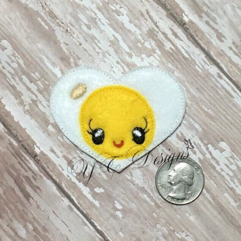 May include: A white felt heart-shaped appliqu&eacute; with a yellow felt yolk inside. The yolk has a smiling face with black eyes and eyelashes.