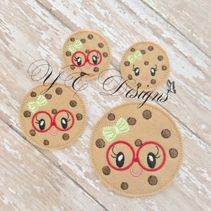 Puede incluir: Tres parches de fieltro bordados con forma de galletas de chocolate con fondo marrón, pepitas de chocolate blanco y un lazo verde. Cada galleta tiene una cara sonriente con gafas rojas y pestañas negras.