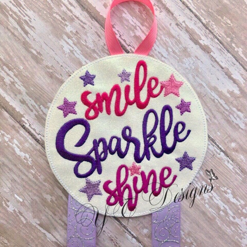 Puede incluir: Un c&iacute;rculo blanco con las palabras "smile sparkle shine" bordadas en rosa, morado y azul. El c&iacute;rculo est&aacute; rodeado de estrellas moradas y rosas. El c&iacute;rculo est&aacute; unido a dos lazos de cinta morada con purpurina plateada.